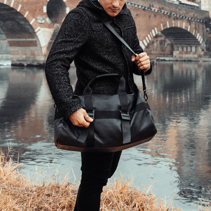 Nordweg black leather bag carried near a bridge and stone wall in a city lifestyle photo