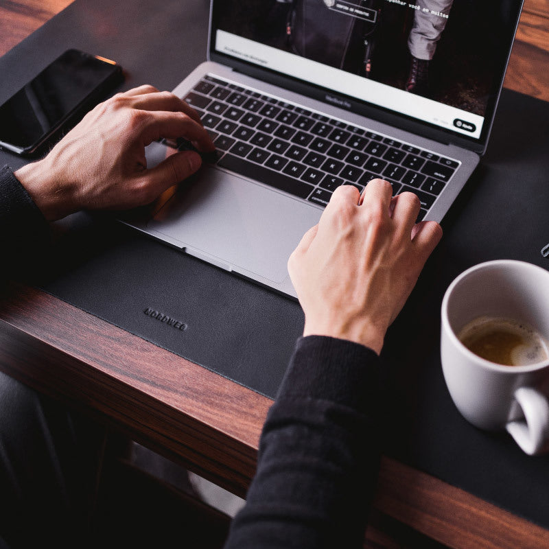 Nordweg black leather desk pad with laptop, hands, and writing tools in a workspace close-up