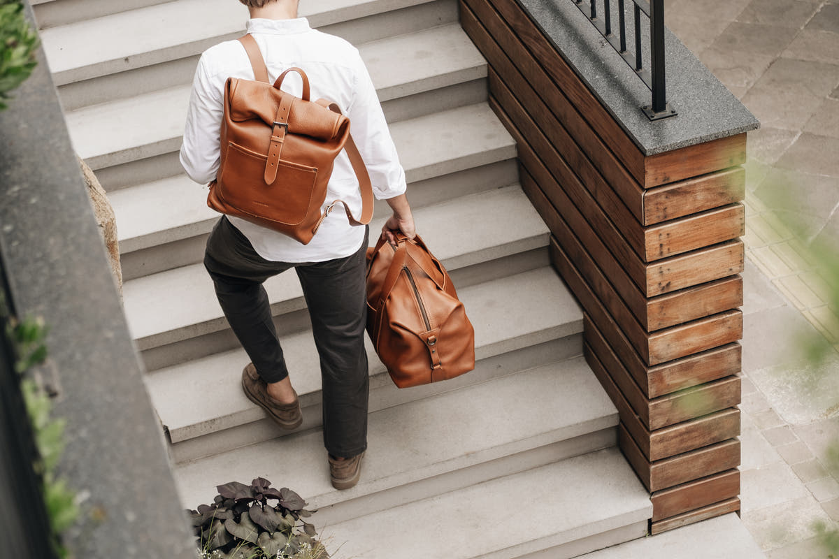 Cognac Nordweg Legacy leather backpack worn while carrying the matching Legacy duffel on stairs, SKUs NWL001.CG and NWL002.CG