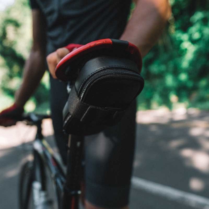 Black Nordweg Serra leather bicycle saddle bag mounted under a red bike seat during a ride, SKU NW090.IP, GTIN 7898571000948