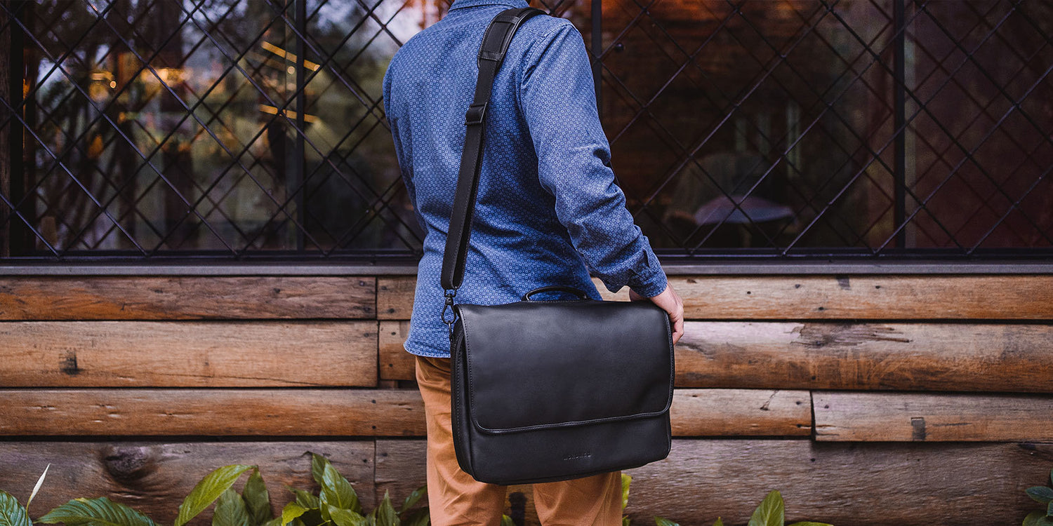 Nordweg black leather messenger briefcase worn over the shoulder beside a wood wall