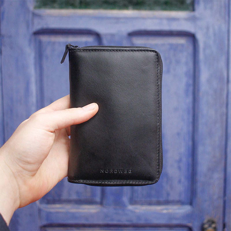 Black Nordweg leather passport wallet held in front of a blue door for the travel essentials collection