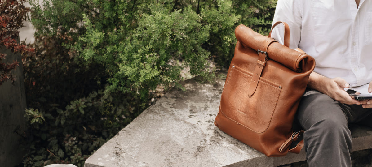 Cognac Nordweg Legacy roll-top leather backpack on a stone bench beside a seated person and green shrubs.