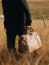 Cognac Nordweg Legacy leather duffel carried low by hand through a grass field, SKU NWL002.CG, GTIN 7898571002300