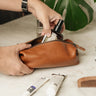 Hand placing a grooming product inside the open Nordweg Legacy Organizer in cognac, with toiletries visible around it