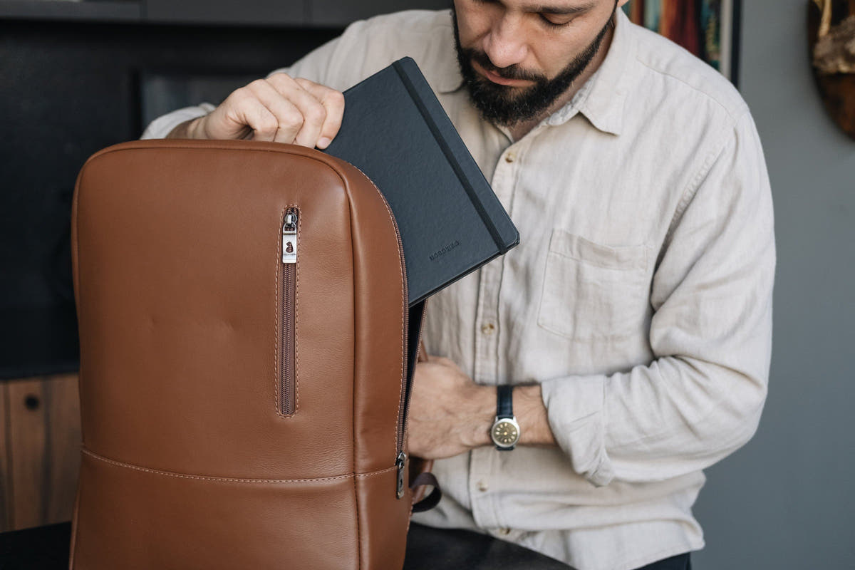 Man placing a black Nordweg notebook inside the caramel Nordweg Nomad leather laptop backpack