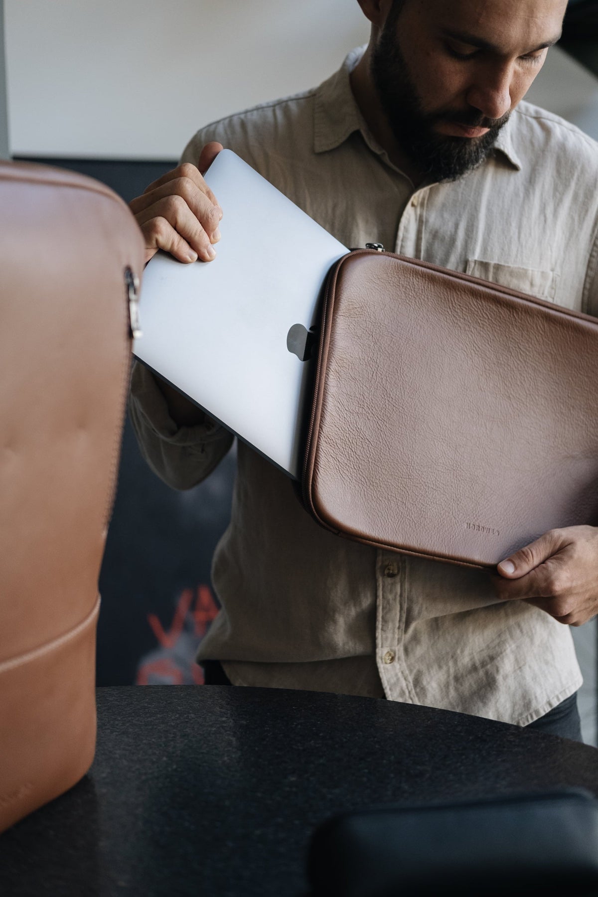 Man placing a laptop into a matching leather sleeve beside the caramel Nordweg Nomad leather laptop backpack