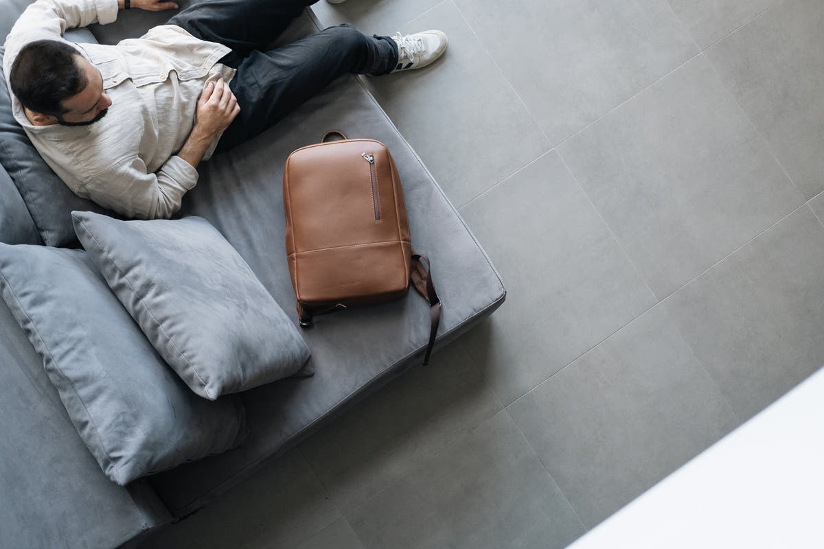 Overhead view of a man and caramel Nordweg Nomad leather backpack beside a gray sofa