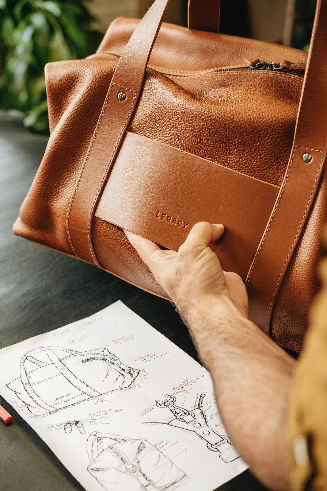 A maker holding a completed leather bag beside the workshop drawings